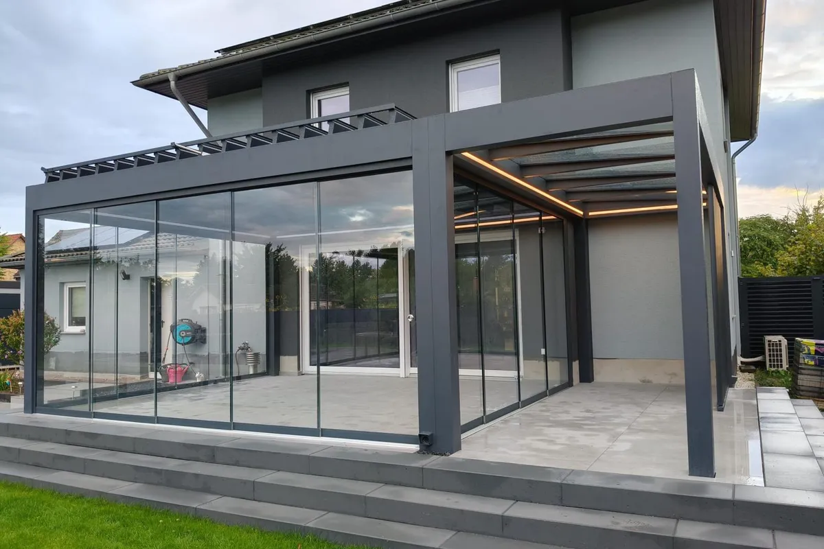 Anthrazitfarbene Pergola mit Glasschiebewänden und LED-Licht in den Trägern an einem Einfamilienhaus