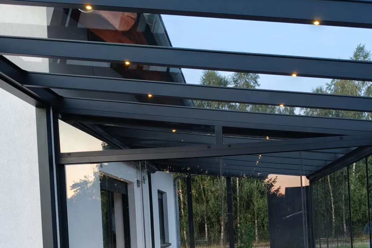 Anthrazitfarbene Pergola mit Glasdach und LED-Spots in den Trägern am Abend