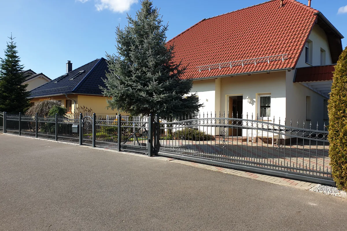 Stahlzaun mit senkrechten Stäben und Schiebetor vor einem Einfamilienhaus mit rotem Dach