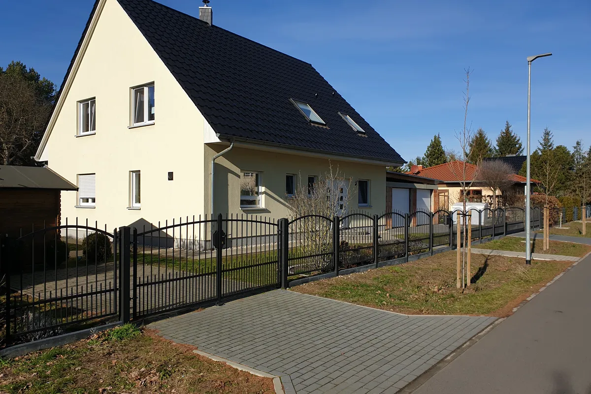 Stahlzaun mit Bogenfeldern und Tor vor einem hellen Einfamilienhaus